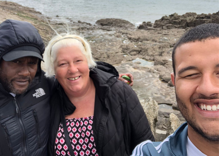 Angela and her family take a selfie at the beach, smiling as they look into the camera. There are rock pools behind them, and the waves lap gently at them in the background.