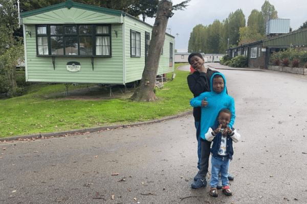 Three children stand on a paved path near a green caravan. One child in a black outfit strikes a pose, another in a blue hoodie gives a thumbs-up, and the smallest child smiles, wearing a vest and jeans. Trees and grass are in the background.