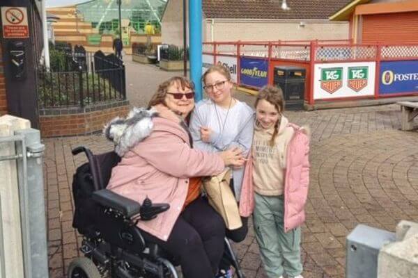  Kay is in her wheelchair at the holiday park whilst her granddaughters, Amelia and Eden, are standing beside her smiling happily.