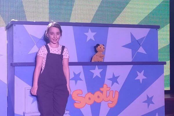 Eden smiling happily and standing in front of a "Sooty" box with Sooty the bear on the box.