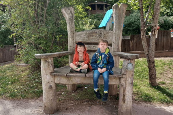 Elijah and Alistair sitting on a giant wood chair in the woods.