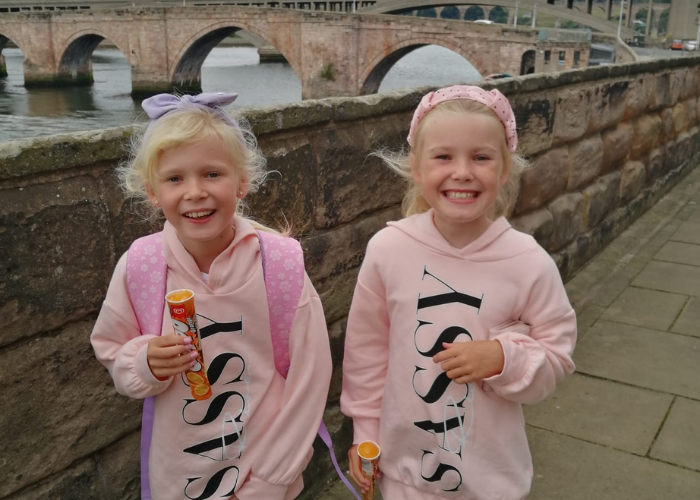 Two sisters wearing matching jumpers stand on an old stone bridge. They both have big smiles on their faces. In the background is a river with another bridge further away. 