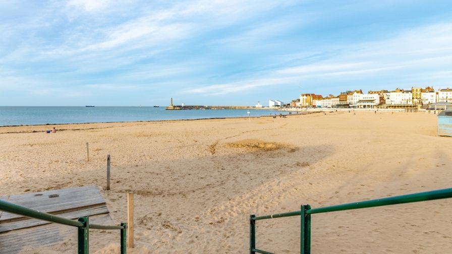 The sun shines on Margate beach with a blue sky above with the sea in view. In the background you can see buildings on the coast.