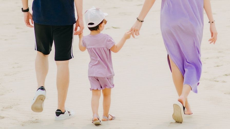 A child and her family walk away across a beach. The child is holding hands with they're Mum and Dad on either side.