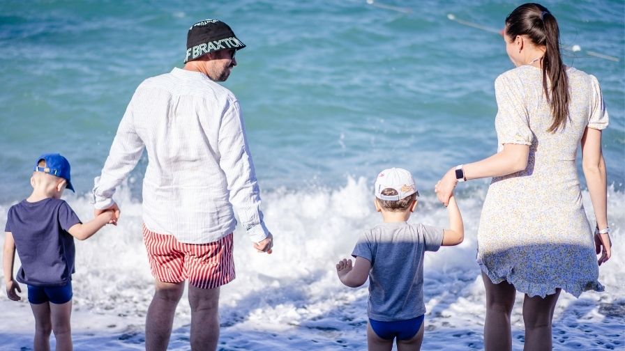 A family; Mum, Dad, Son and Daughter hold hands while standing at the edge of the sea as waves splash forward. 