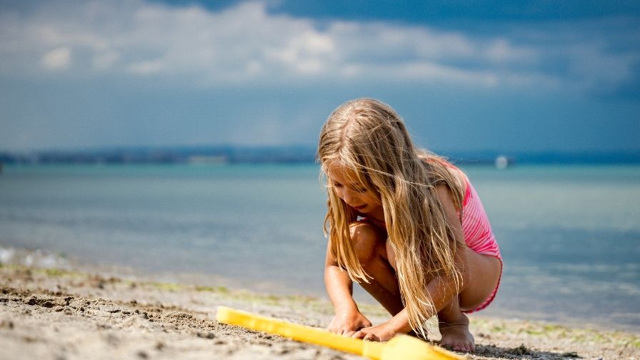 A young girl crouches down while on the beach playing with the sand. Next to her is a yellow plastic spade and the sea lies in the background.