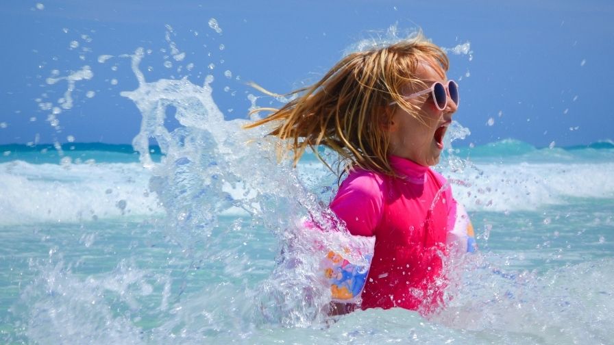 A young girl wearing sunglasses and armbands splashes in the sea. She has a big smile on her face. 