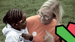 A child and her support worker sit together in a grassy park. They are both looking at each other smiling, and pointing at themselves whilst learning something new.