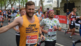 A Family Holiday Charity runner is wearing a bright yellow branded vest as he runs along with a smile on his face, and arm outstretched in greeting to his supporters lining the route out of shot.