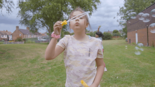 A girl in a patterned t-shirt is outdoors on a grassy field, blowing bubbles with a bubble wand.