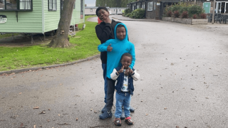 Three children stand on a paved path near a green caravan. One child in a black outfit strikes a pose, another in a blue hoodie gives a thumbs-up, and the smallest child smiles, wearing a vest and jeans. Trees and grass are in the background.