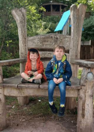 Elijah and Alistair sitting on a giant wood chair in the woods.