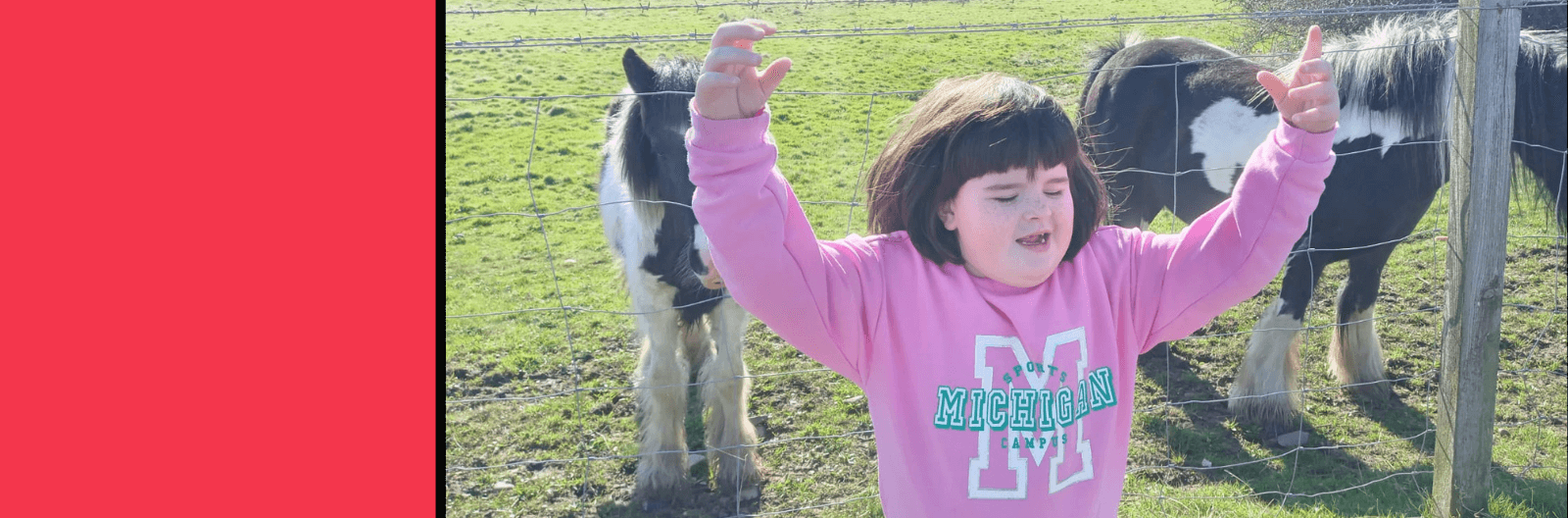 A girl standing in front of a farm holding her hand up smiling happily.