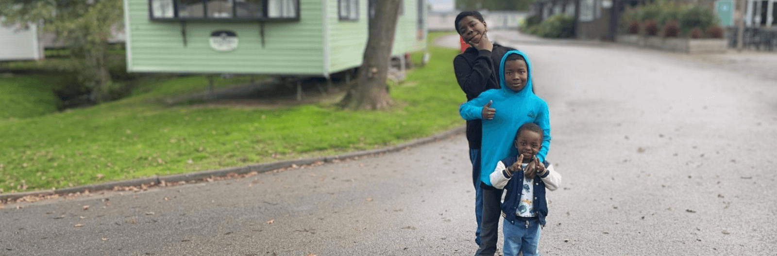 Three children stand on a paved path near a green caravan. One child in a black outfit strikes a pose, another in a blue hoodie gives a thumbs-up, and the smallest child smiles, wearing a vest and jeans. Trees and grass are in the background.