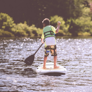 On a sunny and bright day a young boy is on a paddleboard in the water, paddle in hand ready to move forward. In the distance are green bushes and trees.