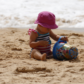 A young toddler sits on the beach she has a swimming costume and apink bucket hat on. She plays with a bucket and sand with the waves rippling in the background. 