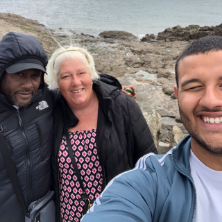 Angela and her family take a selfie at the beach, smiling as they look into the camera. There are rock pools behind them, and the waves lap gently at them in the background.