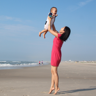 Mum holds her child in the air on the beach on holiday. The sea laps in the background.
