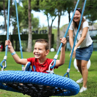 Mum and son play on swings in the playground on holiday. They both have big smiles on their faces, having a lot of fun together.