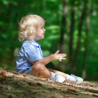 A child sits on the forest floor, clapping his hands in excitement. There are trees behind him as he enjoys his first holiday.