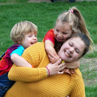 A mum is sat in a green park whilst her 2 daughters are jumping on and giving her big cuddles from behind. Mum is reaching for their arms and the whole family have big smiles on their families, delighted to be in the moment.