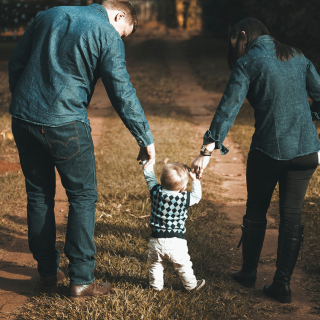 A mum and dad walk a young toddler along a grassy path holding hands.