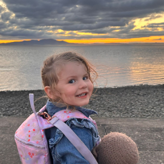 A young girl is standing on the beach with the sun setting over the sea in front of her. Her head is turned towards whoever is holding the camera and she is smiling.