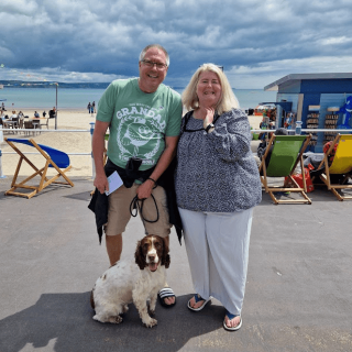 A couple standing in front of the Weymouth Beach with their dog.