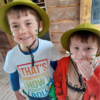 2 young boys wear explorer hats and green lines of face paint on their cheeks whilst smiling.