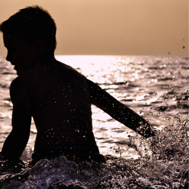 A sunset photo of a boy playing in the sea. The boy is a silhouette but you can see that he's about to make a big splash in the water.