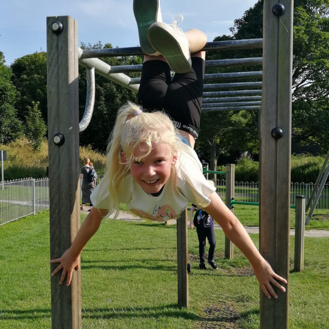 Gillian's granddaughter hangs upside down on a climbing frame. Her legs are looped around the monkey bars, and her arms are outstretched holding the vertical posts on either side. She has a wide grin across her face, and her head is held up toward her grandmother, who is holding the camera and out of shot. 