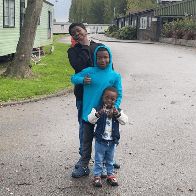  Three children stand on a paved path near a green caravan. One child in a black outfit strikes a pose, another in a blue hoodie gives a thumbs-up, and the smallest child smiles, wearing a vest and jeans. Trees and grass are in the background.