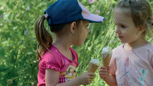 Two young girls are looking at each other and smiling as they hold large ice creams. They're in a park with green trees behind them.