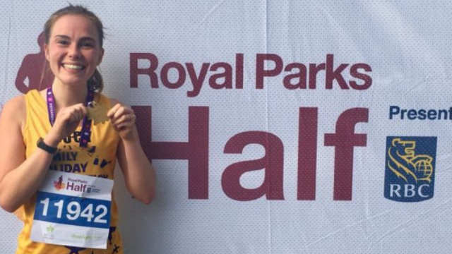 A Family Holiday Charity runner holds her finisher's medal up in celebration with a big smile on her face, next to a Royal Parks Half Marathon banner. 