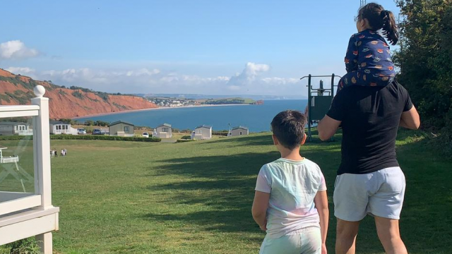 Shania's family stand next to their accommodation from the safety of their holiday park as they look out at a stunning horizon of red cliffs sloping into the bright blue sea below. Shania's daughter is sat on Dad's shoulders, and their son walks alongside